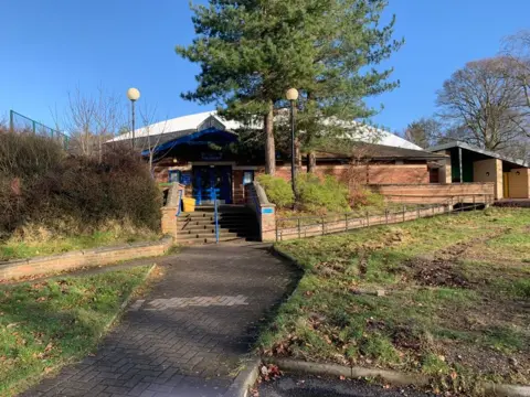 A sports complex building - single story and made of brick with trees in front of it and the curved white roof of the tennis centre behind it