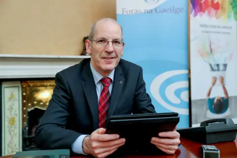 Seán Ó Coinn Seán Ó Coinn in a black suit, blue shirt and red checked tie, holding a tablet in an office with the Foras na Gaeilge backdrop beside a mantlepiece.