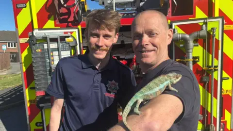 Two fire fighters, one with a chameleon on his arm standing in front of 