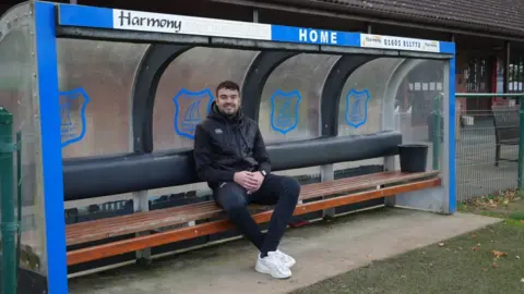 Shaun Whitmore/BBC Wroxham, midfielder Jordan King, wearing black sportswear and smiling. He is sitting on the club bench.