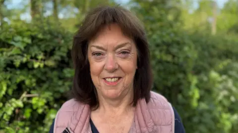 Paul O'Gorman/BBC A middle-aged woman wearing a pink puffer vest stands smiling in front of some bushes.