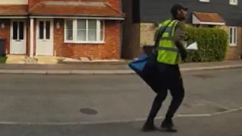 Kent Police A CCTV image of a man wearing a hi-vis jacket and a blue bag across his shoulder. He is on a residential street.