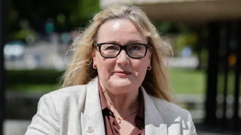 Newcastle City Councilor Karen Kilgour, who has long blonde hair and is wearing black glasses, a gray linen blazer and a brown shirt, smiles at the camera.