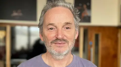 Steve Batts has short hair and a beard. 
He is wearing a purple T-shirt and is standing in a large room with a number of posters on the wall 