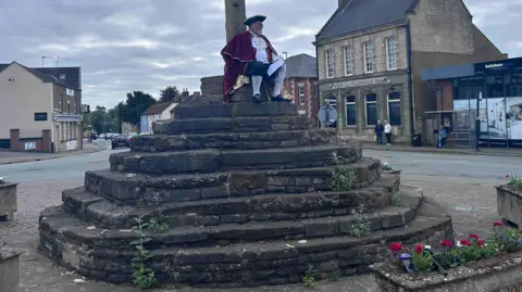 Kate Bradbrook/BBC Stone steps in the centre of a town - there are weeds growing from some steps. A town crier in red and white with a black tricorn hat is sitting on the top step. There are shops and houses in the background, and a stone planter in the foreground.