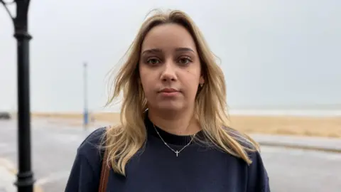 Image shows Viktoriia staring into the camera. She had a blue knitted jumper on. Round her neck, she is wearing a silver cross necklace. She has blond hair that falls just below her shoulder. It is blowing in the wind away from her face. Behind her is Bournemouth beach which is blurred. There is the pavement in the forefront, then the sand and then the sea in the distance. 
