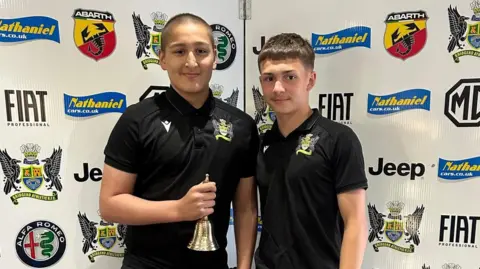 Lloyd Griffiths Ethan in a club rugby shirt with a fellow player. He is holding a brass bell in one hand. 