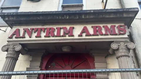 A close up of the front entrance of the former hotel. In the foreground is metal fencing. The portico of the building has decorative stone columns and the door is red with a Georgian style fan light above it. The words Antrim Arms are written in red above the door. The rest of the building is a dirty cream colour. 