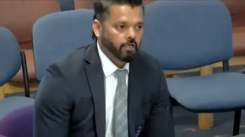 Roy Francis, who has a dark beard and a diamond earring in his right ear, speaking at the BCP Council meeting. He is wearing a dark suit and silver tie. He is sat in front of blue chairs.