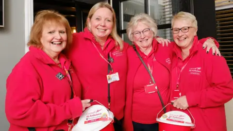Four Devon Air Ambulance volunteers in matching red charity fleeces and polo shirts stood in arms smiling at the camera. Two are holding the charity's donation buckets. 