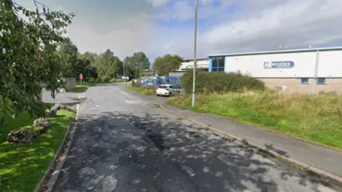 A white industrial unit at Westmorland Business Park in Kendal. There is a worn single track road with potholes skirting around the site. There is green grass and trees on either side of the road.