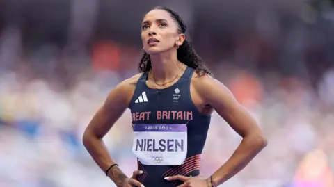 Getty Images Laviai is stood with her hands on her hips looking up. She is wearing a GB navy tight fitting running vest. The words Great Britain are printed in red on her chest. She has a name tag with Nielsen on her front. She has dark brown curly hair tied off her face. 