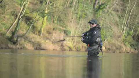 Del Spry standing in a river, the water is up to his thigh. He is casting with a fly fishing rod in his right hand. He is wearing a black jacket, cap and sunglasses on his head and a khaki-coloured chest pack with fishing gear attached to it. The riverbank on the opposite side is lined with trees.