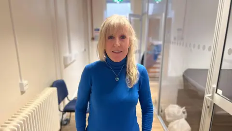 A woman with blonde hair wearing a blue turtle neck jumper stands in a hallway