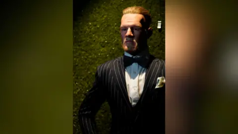 National Wax Museum Plus Patung lilin Conor McGregor dalam setelan jas berdiri di depan latar belakang hijau.