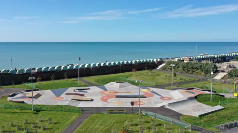 Maverick Skateparks A skatepark by Maverick Skateparks at Hove Beach Plaza near Brighton. It is large in size and is surrounded by grass. The sea can be seen behind it.