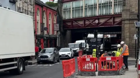 BBC A busy road with a railway bridge overhead and men in orange jackets and white protective helmets by the roadside