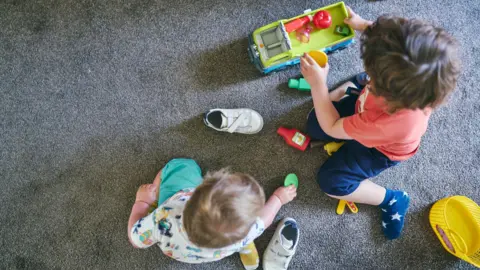 Getty Images As seen from above, two children are sat on a grey carpet. One has fair hair and is wearing green shorts and a white printed top. Their shoes are next to them and they are playing with a green disc. One has dark hair and is wearing an orange top and navy shorts and socks. They are playing with a truck with plasic food items in it.