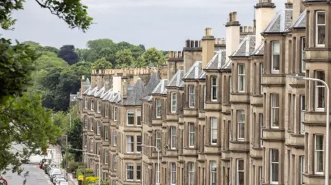A row of closely packed, beige, stone-terraced houses with bay windows and tall chimneys line a slightly sloping residential street. Parked cars run along the left side of the road, while a leafy tree branch frames the scene in the foreground