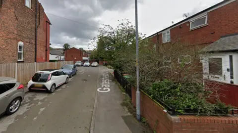 Google Google Streetview of Gateshead Close in the Rusholme area of Manchester. There is a row of parked cars on one side, and several red-brick houses on both sides and at the end of the close.