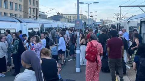 A large crowd of people standing on the platform at Cambridge railway station. There are trains parked either side of the platform.