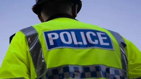 The back of a police officer taken from a low angle. The male officer is wearing a yellow hi-vis jacket with silver reflective strips and the word "POLICE" on it. The officer is wearing a traditional hard police hat. The sky in front of him is blue.