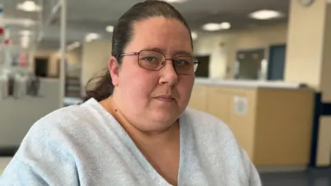 A woman with dark brown hair tied back, glasses and a grey V-neck top is sat down lookkng at the camera. There is a room with strip lighting and some furniture behind her.