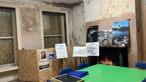 Photograph of one of the rooms inside Rock Hall. The image includes a desk, an old fireplace and boarded-up windows.
