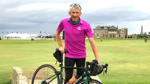 Thea Lawson The image shows Peter Lawson standing next to his push bicycle, he's wearing black trousers, a bright pink t-shirt and has sunglasses resting on top of his head. There is green grass and a historic looking building behind him.