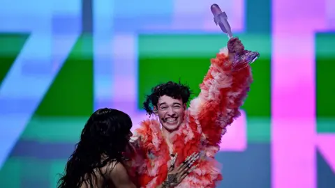 loreen and nemo embrace as nemo holds the eurovision song contest trophy aloft. loreen has long black curly hair and long silver nails, nemo is wearing a pink and red ruffled coat