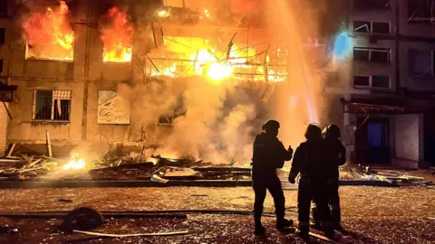 Three firefighters in silhouette fight a blaze in a damaged building. 