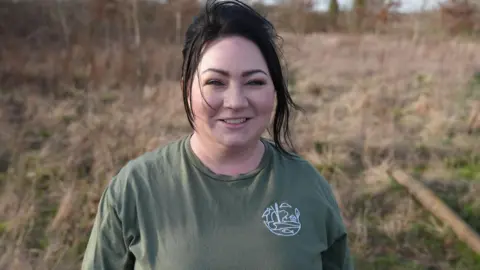 A woman wearing an olive‑green T‑shirt with a small nature‑themed logo on the chest stands in an open, grassy field with dry, pale vegetation stretching into the background. Their dark hair is tied back, blowing slightly in the breeze.