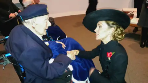 Chris Redston A photo of an old man sitting in a wheelchair wearing a military cap. The Princess of Wales crouches down to shake his hand, wearing a black jacket and a poppy.