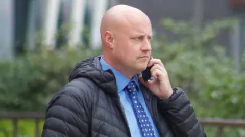 A man with a shaved head walks outside with a mobile phone to his ear