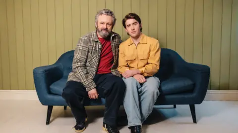 Danny Kaan/ Margo Jones Agency Sheen and Howells sitting next to each other on a navy sofa, looking at the camera. Sheen, on the left, wears navy jeans, a red t-shirt and a brown tartan jacket. Howells wears a yellow jacket, blue jeans and black boots. 