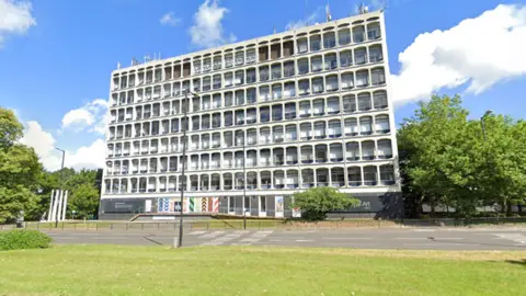 Google is a high, cruelist building on the side of a main road. The sky in the background is bright blue and has trees and grass around the building. There are several masts at the top of the building.