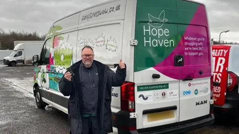 Phil Stout holding a set of keys and giving a thumbs-up gesture as he stands in front of the "wheels of wellness" van. He has short dark hair, a grey beard and glasses. The white van is decorated with the charity's name and colourful graphics.