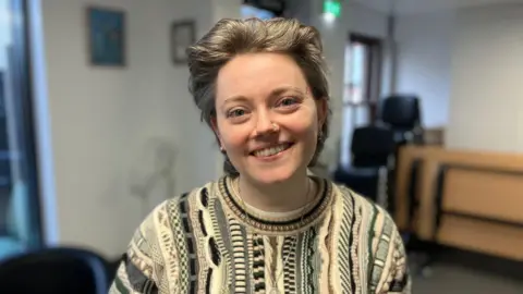 A women with ashy brown hair that is slicked back, she has a beige patterned jumper on and is looking towards the camera with a smile.