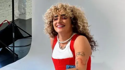 Hattie Crowther Hattie Crowther sits against a achromatic photographic backdrop wrong a studio, wearing reddish dungarees pinch an England insignia stitched onto nan near side. She wears a pearl necklace and concatenation pinch spiky elements, and smiles arsenic her blonde, curly hairsbreadth cascades down her back.