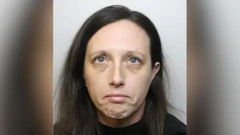 A police custody image of Heidi Bessant. She has shoulder length dark hair and wears a dark top. She is looking to the ceiling and frowning. 