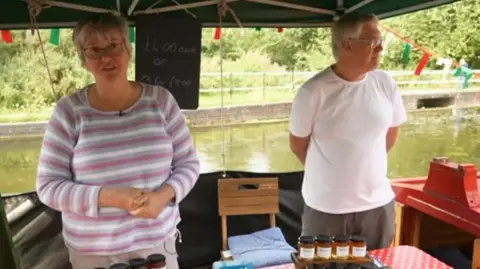 A woman in a stripy pink, white and grey jumper with a man in a white t-shirt selling jam from a canal boat