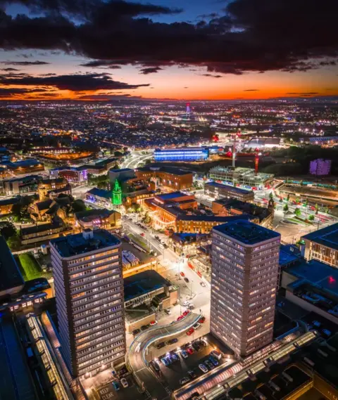 Ross Johnston Aerial view of two tower blocks and lit city streets, with an orange sky and dark clouds in the background.