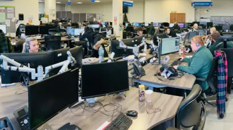 Rows of wooden desks facing each other with two screens on each. People wearing dark green uniforms and headsets sit at most of the desks. A large, open-plan office can be seen behind them.