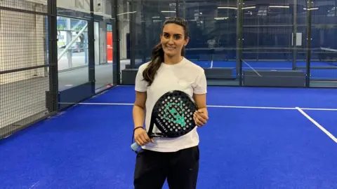 BBC Kiranjit is standing on a blue-floored padel court. The glass walls and black railings of the court can be seen in the background. She is holding a black and blue padel racket in both hands. Kiranjit is wearing a white T-shirt and black leggings. Her long black hair had been tied back into a high ponytail and she is smiling with teeth into the camera.