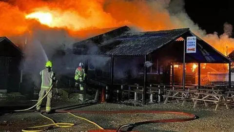 Fire fighters tackling a blaze at Mrs T's fish and chip shop. One has a water hose on the building, 