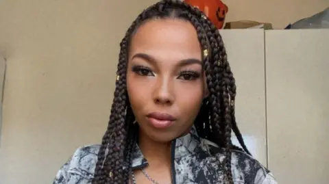 Kelisha Walker The person with braids is wearing a patterned jacket over a black top, accessorised with layered chains. Behind them, kitchen cupboards and household items are visible in the background.
