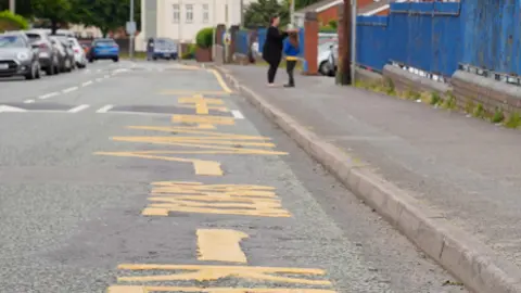 There are new road markings outside a school. Large yellow letters are painted on the road and several cars are parked on the left-hand side.