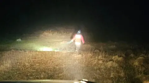 A man is seen from behind walking across moorland with a flashlight illuminating the ground to the side and front of him. He is wearing an orange high-viz jacket 