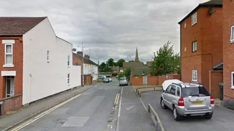 Gladstone Street, Raunds, showing a road down to terraced houses on the left and a block of red-brick flats on the right. There are various cars parked.