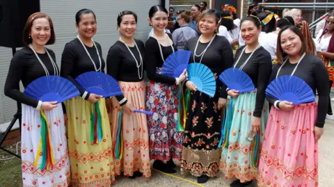 Luton & Dunstable University Hospital Seven nurses holding blue fans, smiling and looking at the camera. They all have a black top on with pearl necklaces on and colourful skirts. They are standing by a grey building. 